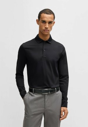 Polo noir à manches longues avec un col moyen et trois boutons, associé à un pantalon gris clair et une ceinture noire avec une boucle en argent.