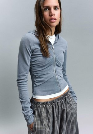 Fiatal nő hosszú barna hajjal, szürke, cipzáras, testhezálló kapucnis pulóvert visel fehér felső fölött, és laza, fekete-fehér kockás nadrágot.