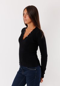 Maglietta nera a maniche lunghe a coste con scollatura a V e dettaglio a catena sul collo. Abbinata a jeans attillati blu scuro.
