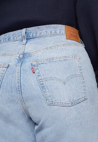 Jasnoniebieskie dżinsy Levi's 501 noszone z ciemnonawowym topem, widoczne tylne kieszenie, szwy oraz skórzana metka marki na pasku.