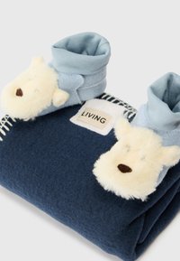 Pătură din fleece albastru cu botine în formă de ursuleț crem. Botinele au fețe moi din blană artificială și vârfuri răsucite. Eticheta spune "LIVING."