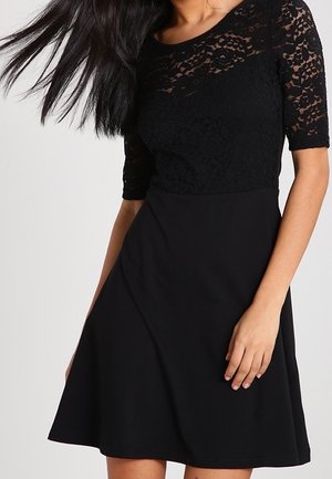 Mujer con vestido negro con mangas de encaje floral hasta la mitad y una falda lisa acampanada, con el cabello largo y oscuro cayendo hacia un lado.