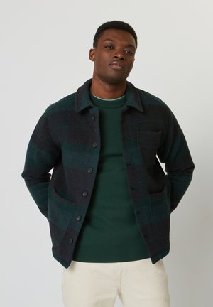 Veste en mélange de laine avec des rayures vert foncé et marine, fermeture à boutons, col et deux poches avant, portée sur un pull vert.