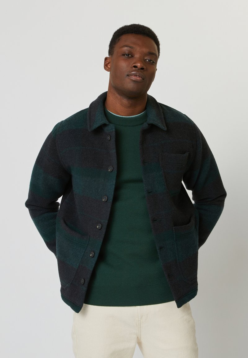 Chaqueta de mezcla de lana con rayas verde oscuro y azul marino, con cierre de botones, un cuello y dos bolsillos frontales, llevada sobre un suéter verde.