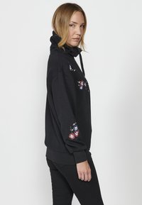 Sweat à capuche noir avec un col haut, orné de broderies florales rouges, bleues et blanches sur les épaules et les manches. Tissu texturé et coupe décontractée.