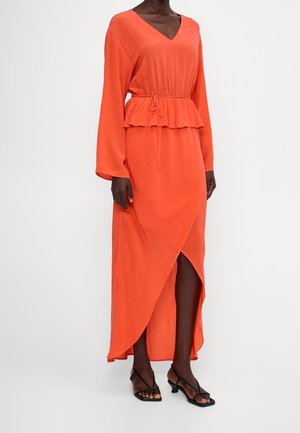 Femme portant une longue robe portefeuille orange à manches longues, taille cintrée, et des sandales noires à lanières à talons bas, debout sur un sol blanc.