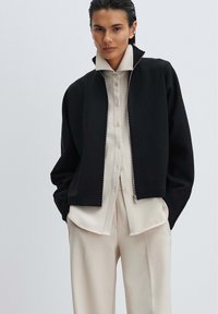 Black cropped jacket with a zippered front, worn over a light beige button-up shirt and loose beige trousers. Smooth fabric with a modern fit.