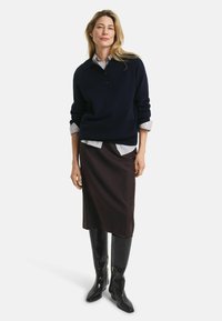 Donna in piedi con un maglione blu navy sopra una camicia a righe, una gonna midi scura e stivali neri alti fino al ginocchio con texture, su uno sfondo neutro.