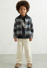 Tamprinė striukė tamsiai mėlynos, pilkos ir žalios spalvų atspalvių, juoda grafikos marškinėliai, kreminės spalvos kargo kelnės ir rudos spalvos šlepetės. Textūruoti plaukai.