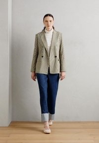 Blazer à double boutonnage à carreaux dans des nuances de vert clair et foncé, porté sur un col roulé blanc, associé à un jean taille haute et des chaussures.