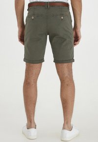 Olivenfargede bomullsshorts med oppbrettede ermer, to baklommer med knapper, og et brunt lærbelte. Hvite sneakers fullfører antrekket.