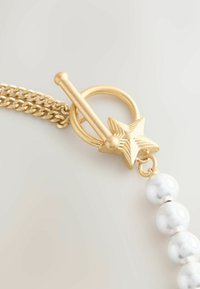 Zlatý kovový otočný uzáver v tvare hviezdy, pripevnený na reťaz s hladkými bielymi koralikmi. Koraliky majú mramorovú textúru.