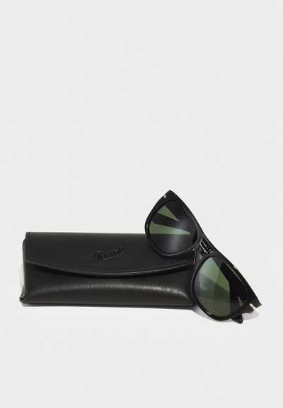 Occhiali da sole neri con lenti verdi appoggiati su un astuccio in pelle nera, caratterizzato da una texture liscia e logo del marchio impresso.