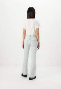 Jean évasé bleu clair avec une texture délavée, design à cinq poches et un patch en cuir au dos. Associé à un t-shirt blanc uni et des chaussures noires.