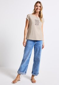T-shirt beige à manches courtes avec impression textuelle, associé à un jean large bleu clair retroussé aux poignets, et des sandales beiges à fines lanières.