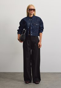 Donna che indossa una giacca di jeans corta blu scuro, pantaloni neri a gamba larga, tiene una borsa nera e grandi occhiali da sole, in piedi su un pavimento grigio.