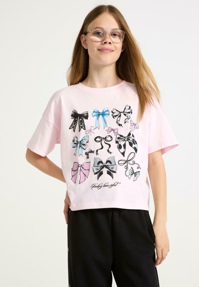 Lyserød t-shirt med korte ærmer, der har et grafisk tryk af forskellige sløjfedesigns i sort, blå og lyserød, med teksten "Feeling bow-tiful" nederst.