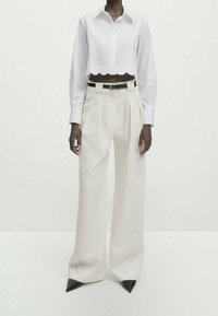 Chemise blanche courte avec ourlet festonné, associée à un pantalon crème taille haute avec plis. Ceinture noire avec accessoires en argent.