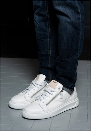 White leather sneakers with side zippers and laces worn with dark blue rolled-up jeans on gray tiled floor.