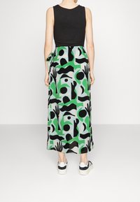 Maxi rok met een abstract patroon in groen, zwart en lichtblauw, met een strikdetail in de taille en gecombineerd met zwart-witte sneakers.