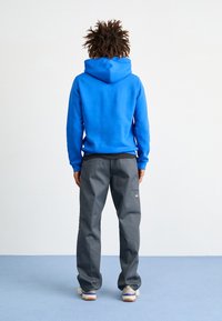 Sudadera azul con un bolsillo tipo canguro y capucha con cordón, combinada con pantalones grises que tienen un bolsillo lateral y zapatillas color beige.