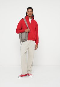 Pulover roșu din fleece cu fermoar, pantaloni largi crem și adidași roșii. Se poartă o geantă gri tip acoperiș cu o suprafață texturată.