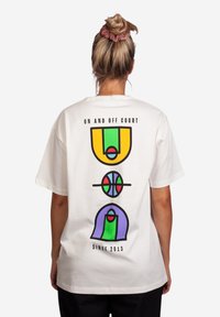 T-shirt bianca a maniche corte con stampa grafica colorata: campi da basket verdi e gialli, palloni da basket rossi e blu e accenti viola.
