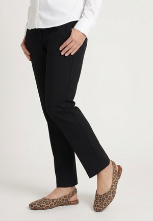 Persoon die zwarte smalle broek, wit overhemd en leopardprint slingback platte schoenen draagt, staand met een been licht opgetild.