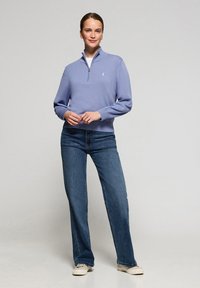 Ljusblå tröja med dragkedja upptill, tillverkad av mjukt stickat tyg, kombinerad med mörkblå jeans med raka ben och vita sneakers.