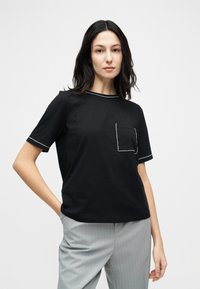 Γυναίκα με σκούρα μαλλιά, φορώντας μαύρο μπλουζάκι με κοντά μανίκια και λευκά ραφές, καθώς και ανοιχτό γκρι παντελόνι με λεπτές ρίγες, ποζάρει με το ένα χέρι στην τσέπη.