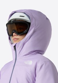 Μωβ αδιάβροχο μπουφάν με ψηλό γιακά και μεγάλο κουκούλι· διαθέτει πλήρες φερμουάρ, λογότυπο στο στήθος και υφή στο φινίρισμα.