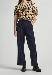 Donna che indossa un maglione a fantasia beige e marrone, pantaloni larghi a righe blu navy e stivali neri stringati, in piedi con una mano in tasca.