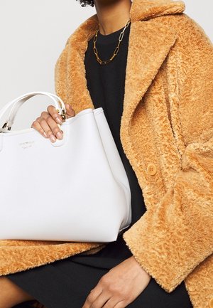 Borsa a mano in pelle bianca con superficie texture e dettagli in oro, tenuta contro un cappotto arancione soffice. Design semplice e moderno.