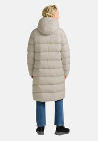 Beige pufferjack met capuchon, voorzien van horizontaal gestikte secties, knielange snit en elastische manchetten. Gedirigeerd over blauwe jeans en laarzen.