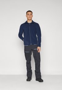 Fleece jacket in navy with front zipper, paired with dark pants featuring multiple zippered pockets and blue accents.