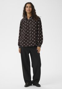 Rjava srajca z gumbi in belimi pikami, z dolgimi rokavi z gumbi na manšetah, kombinirana z ohlapnimi črnimi hlačami in črnimi čevlji.