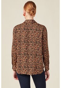 Blus med leopardprint i brunt, svart och rött; har krage, långa ärmar och en slät tygstruktur. Knappdetalj vid ärmsluten.
