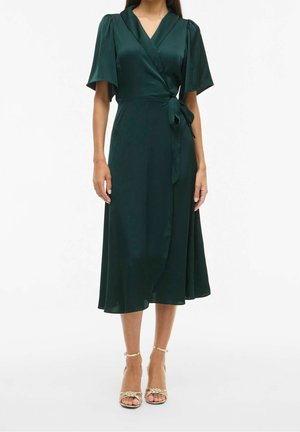 Femme portant une robe midi portefeuille vert foncé avec des manches courtes bouffantes et des sandales dorées à talons hauts, debout sur un fond blanc.