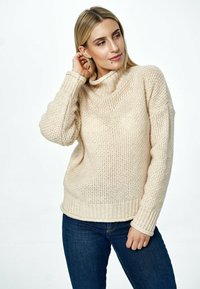 Beżowy sweter dziany z golfem, �ściągaczem na dole i szerokimi rękawami. Tekstura jest luźna i gruba, co nadaje mu przytulny wygląd.