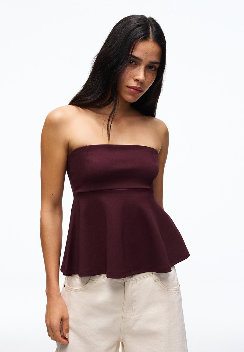 Jeune femme aux longs cheveux foncés portant un top bustier bordeaux foncé à basque et un pantalon beige clair, posant sur fond blanc.