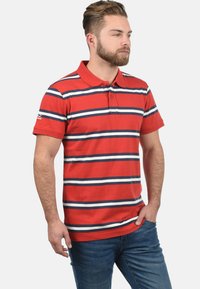 Rotes Poloshirt mit marineblauen und weißen horizontalen Streifen, mit Kragen und kurzen Ärmeln, kombiniert mit blauen Jeans.