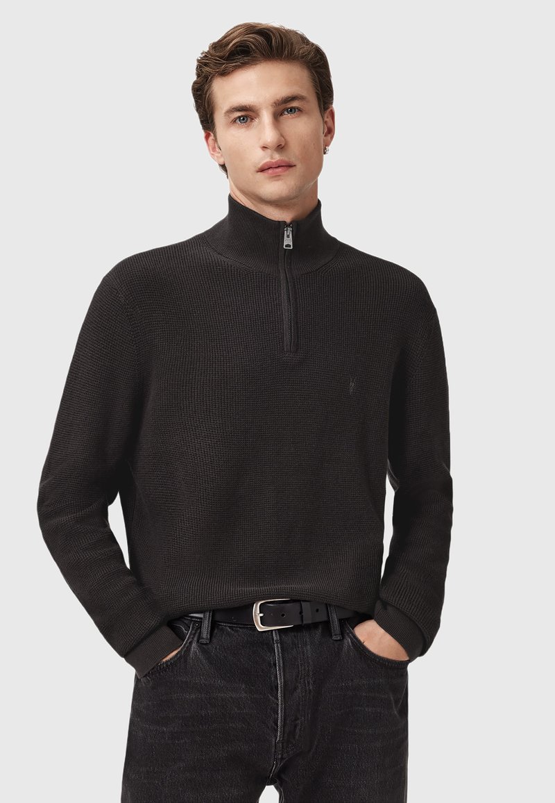 Homme à la peau claire et aux cheveux bruns courts portant un pull noir à demi-zip et un jean noir, les mains dans les poches, sur fond uni.