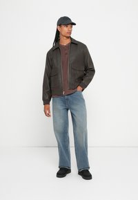 Mies, joka pitää päässään mustaa lippistä, tummanruskeaa nahkatakkia, ruskeaa henley-paitaa, vaalean sinisiä farkkuja ja mustia kenkiä, seisoo valkoista taustaa vasten.