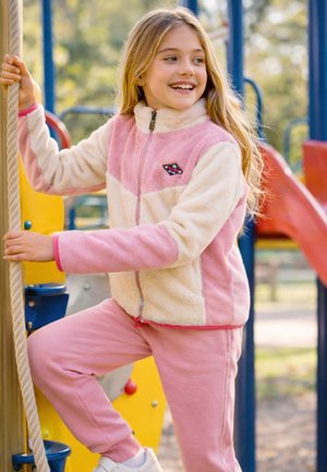 Jeune fille portant une veste polaire rose et crème grimpant à une corde sur une aire de jeux colorée, souriante et regardant sur le côté.