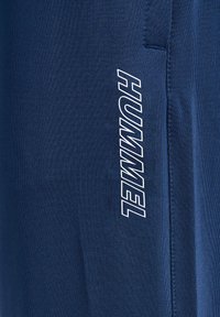 Jūras spēku sporta bikses, kas izgatavotas no teksturēta auduma, ar baltu "HUMMEL" logo sānos un sānu kabatas detaļu.