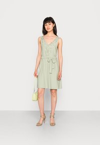 Vestido sem mangas verde-claro com decote em V, bordados florais e cintura com laço. Combinado com uma mala a condizer e saltos finos.