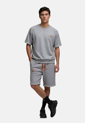 Homme debout, les mains dans les poches, portant un sweat-shirt gris à manches courtes et un short assorti avec des cordons orange et des chaussures noires.