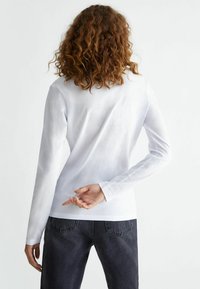 White long-sleeve shirt viewed from the back, highlighting smooth fabric, a fitted design, and a plain hem. Paired with dark jeans.