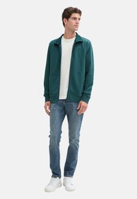 Giacca zip in teal con due tasche frontali, indossata sopra una maglietta bianca, abbinata a jeans azzurri chiari e sneakers bianche.