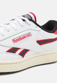 Baskets en cuir blanc avec accents rouges et noirs, dotées d'une semelle en caoutchouc texturée et du logo Reebok sur le côté. Fermeture à lacets.